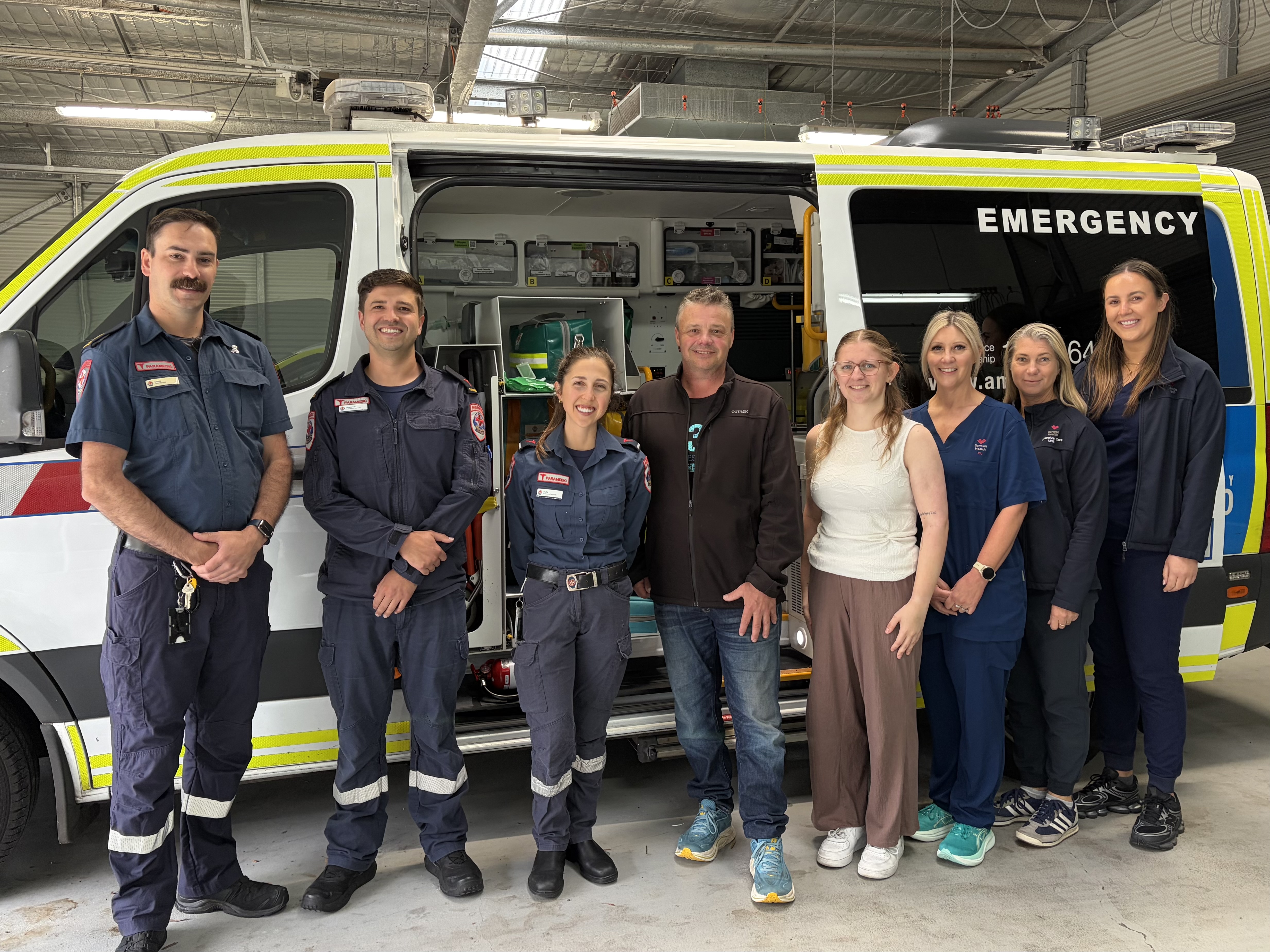 Paramedics, nurses and a man and young woman in casual clothes smile for the camera in front of an ambulance.