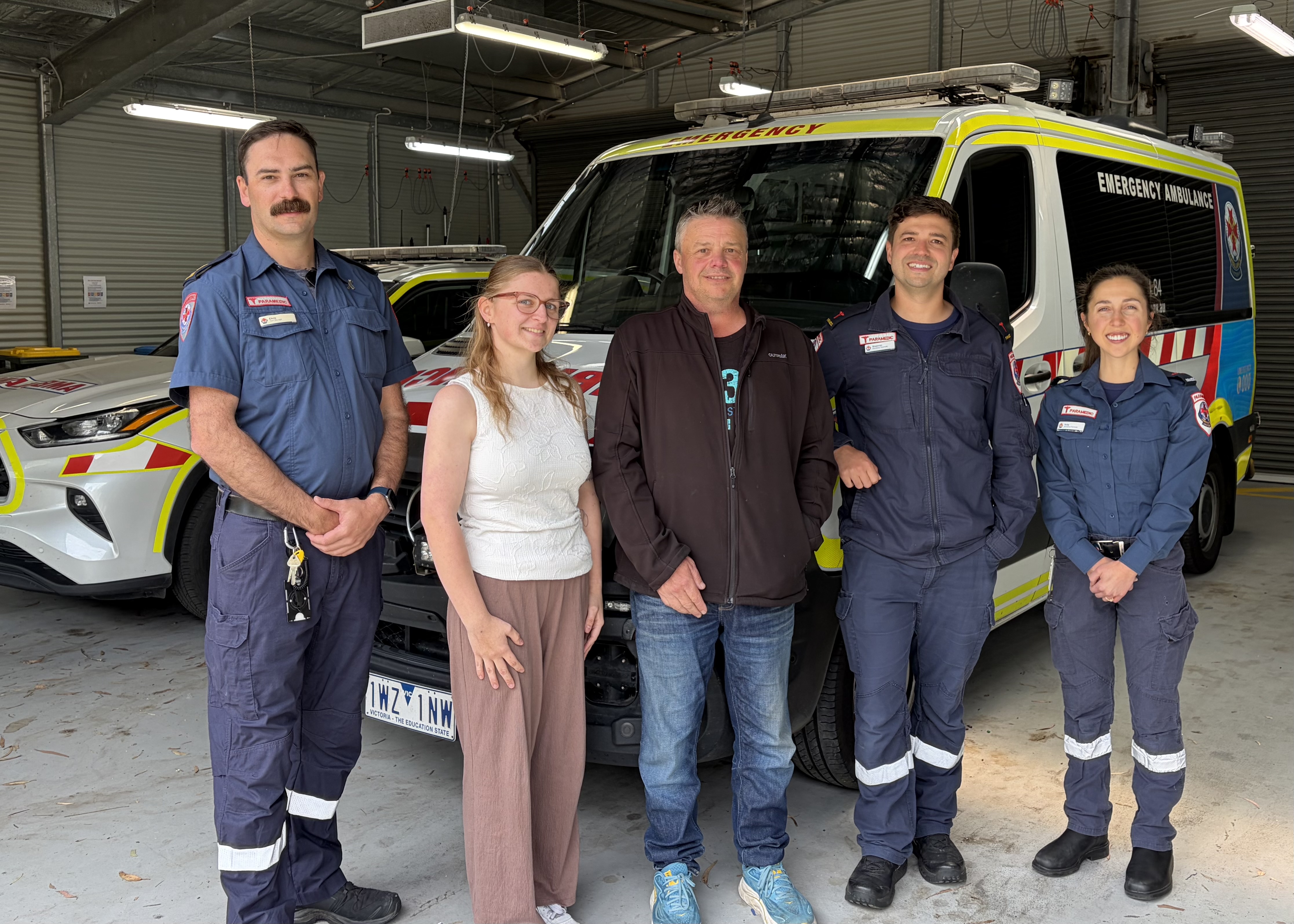 A man and a young woman in casual clothes smile for the camera with three paramedics in front of an ambulance.