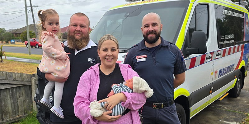 Paramedic Joel Woolley standing with Todd Gardner and Cassie Walters in front of an ambulance. Todd and Cassie are holding their children Elliotte Gardner and baby Levi in their arms.