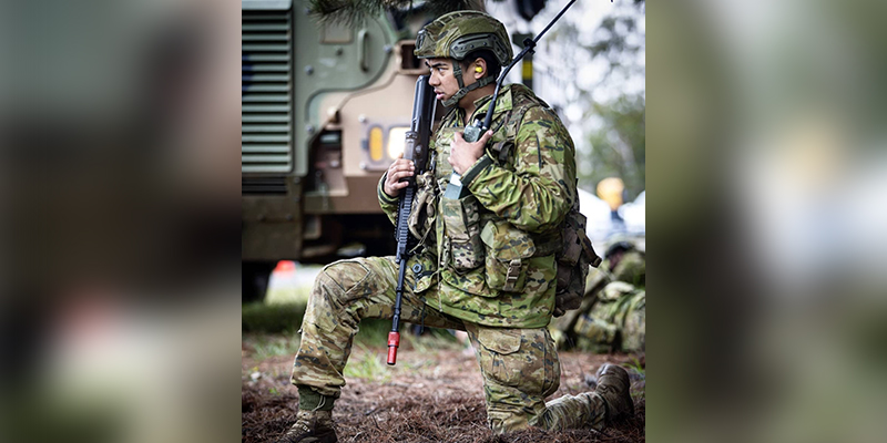Paramedic and Army Reserve Rifleman Josh Pineda in full army gear kneeling and communicating via a radio that is attached on his chest.