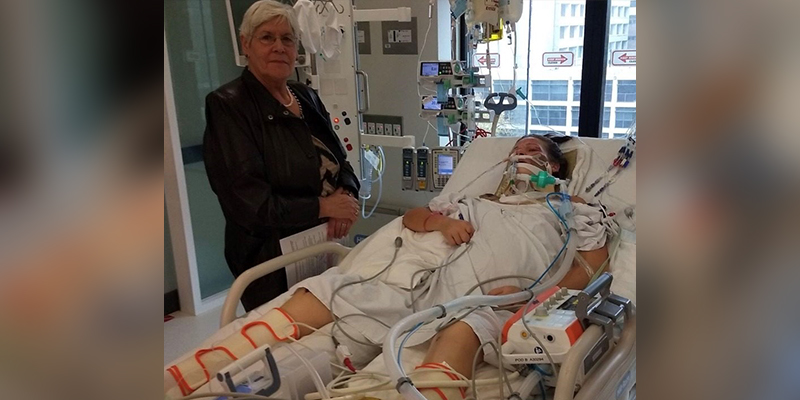 Astrid's worried mother standing beside Astrid while she is resting on the hospital bed. Astrid is surrounded with hospital equipment and support tubes aiding her with her recovery.