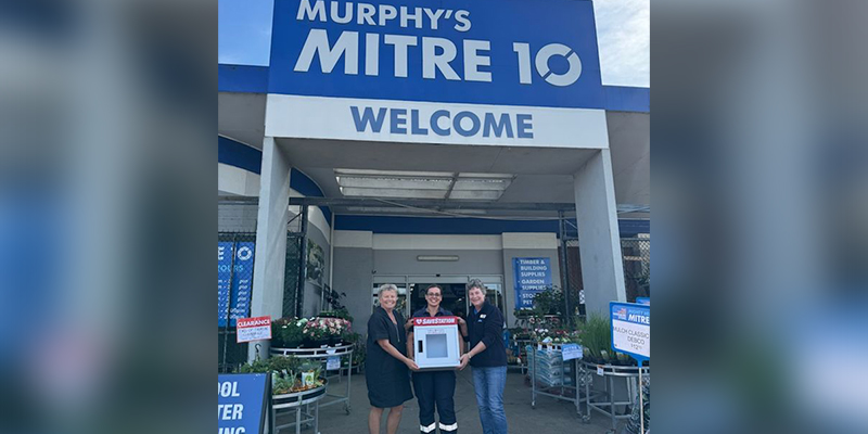 Three ladies standing in front of a Mitre 10 store while holding the cabinet that houses the automated external defibrillator.