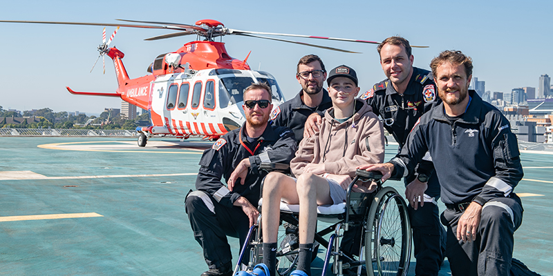15-year-old James is seated in a wheelchair and in front of the Ambulance Victoria helicopter. Four Ambulance Victoria flight paramedics are beside James.
