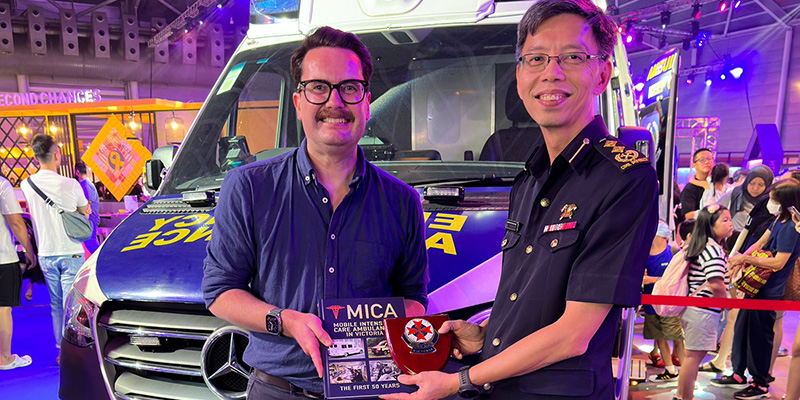 Ambulance Victoria Medical Director Associate Professor David Anderson presenting the Mobile Intensive Care Ambulance (MICA) book to SCDF Director of Emergency Medical Services Department Senior Assistant Commissioner Yong Meng Wah in Singapore in 2023. Both men were standing in front of an ambulance in an exhibition hall.