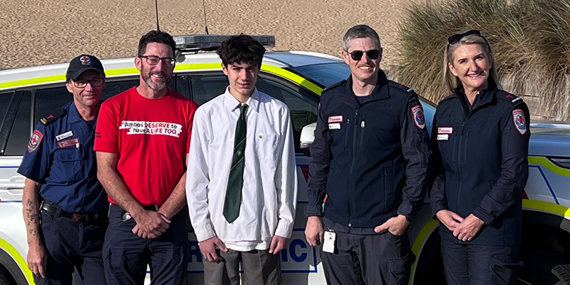 Giordi standing together with the four paramedics that came to his aid. All five are standing beside an Ambulance Victoria vehicle that is parked beside the beach.
