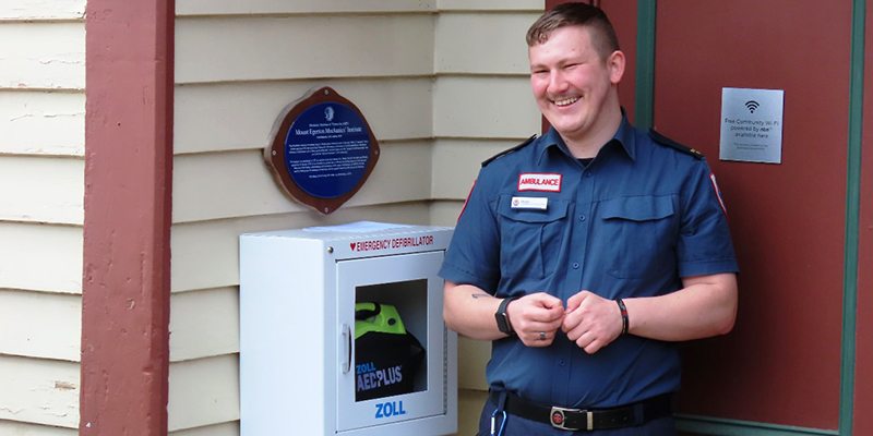 Ambulance Community Officer Brodie standing beside the newly installed automated external defibrillator at Mount Egerton Mechanics Hall.