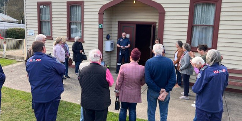 A group of about 15 locals standing around Ambulance Community Officer ACO and listening to his presentation about how to use the automated external defibrillator.