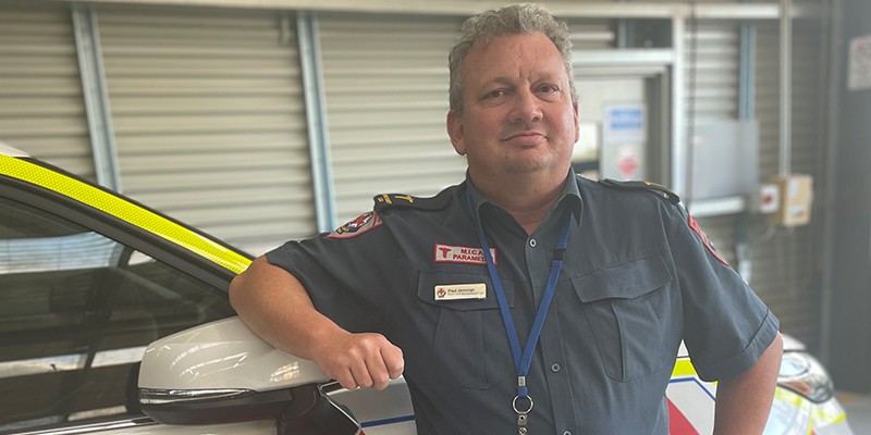 Paul is standing proudly while resting one arm on an Ambulance Victoria vehicle's side mirror.